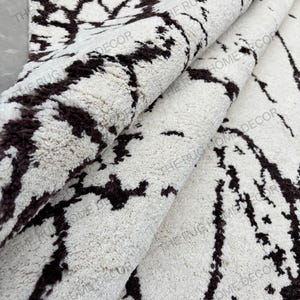 May include: A rolled-up, off-white rug with a dark brown, abstract pattern. The rug has a thick, textured pile and is partially rolled, revealing the underside. The words "THE RUG HOME DECOR" are printed on the rug.