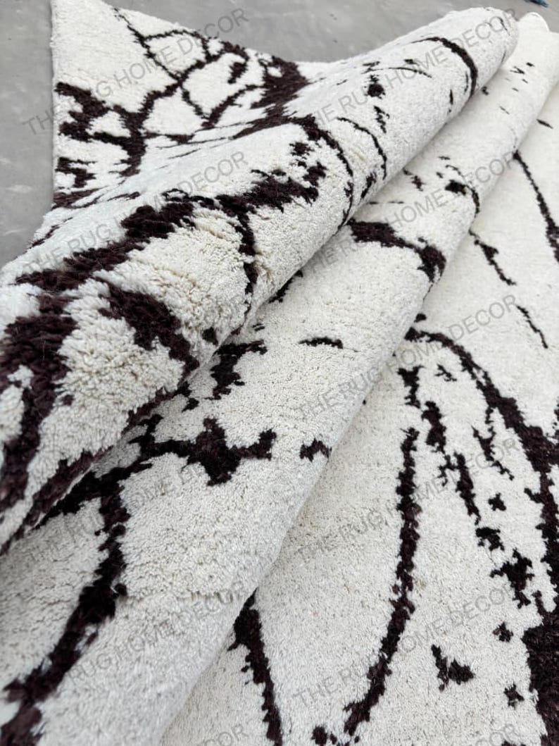 May include: A rolled-up, off-white rug with a dark brown, abstract pattern. The rug has a thick, textured pile and is partially rolled, revealing the underside. The words "THE RUG HOME DECOR" are printed on the rug.