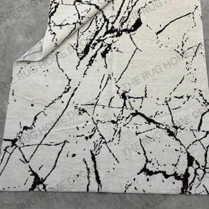 May include: A rectangular rug with a white background and a black marble-like pattern. The rug has a folded corner, revealing the underside. The words "THE RUG HOME DECOR" are printed on the rug.