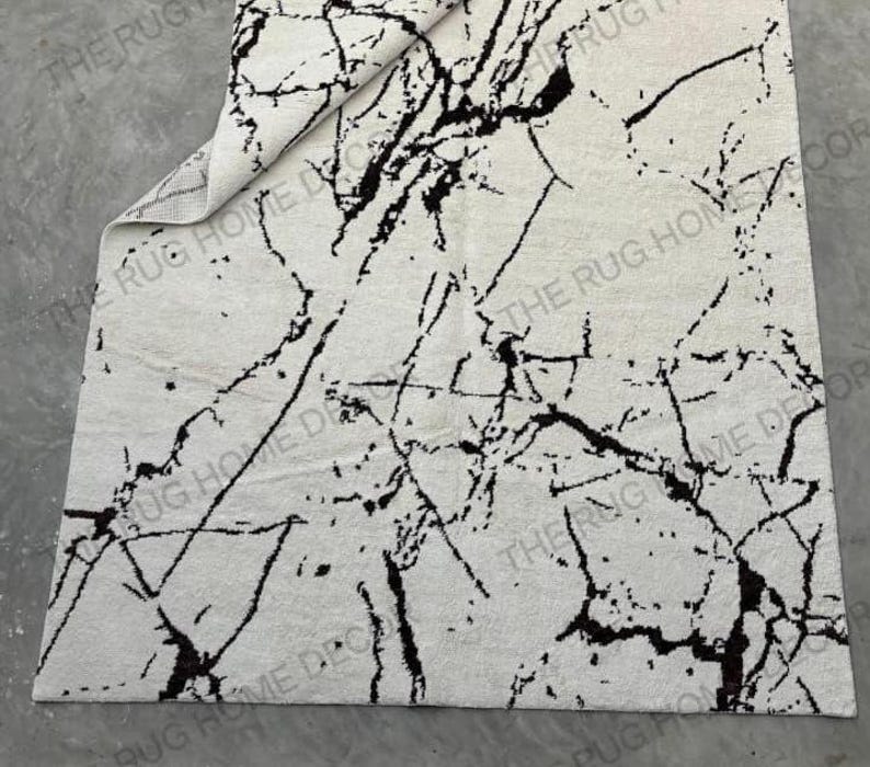 May include: A rectangular rug with a white background and a black marble-like pattern. The rug has a folded corner, revealing the underside. The words "THE RUG HOME DECOR" are printed on the rug.
