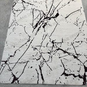 May include: A rectangular, off-white rug with a black, marble-like pattern. The rug's design features dark, irregular lines that create a striking contrast against the lighter background. The rug is a home decor item.