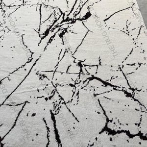 May include: A rectangular white rug with a black marble pattern. The rug's design features irregular black lines and veins across the white surface, creating a natural stone appearance. The rug is a home decor item.