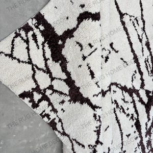 May include: A close-up of a white rug with a dark brown, abstract pattern. The rug has a soft, textured surface and a neutral color palette. The words "THE RUG HOME DECOR" are printed on the rug.