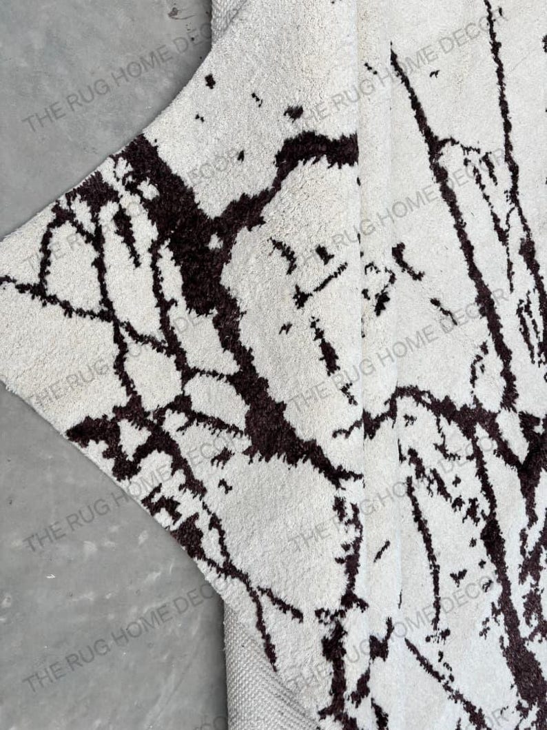 May include: A close-up of a white rug with a dark brown, abstract pattern. The rug has a soft, textured surface and a neutral color palette. The words "THE RUG HOME DECOR" are printed on the rug.