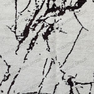 May include: A close-up of a white rug with a black abstract design. The rug has a textured surface and the words "THE RUG HOME DECOR" are printed on the rug.