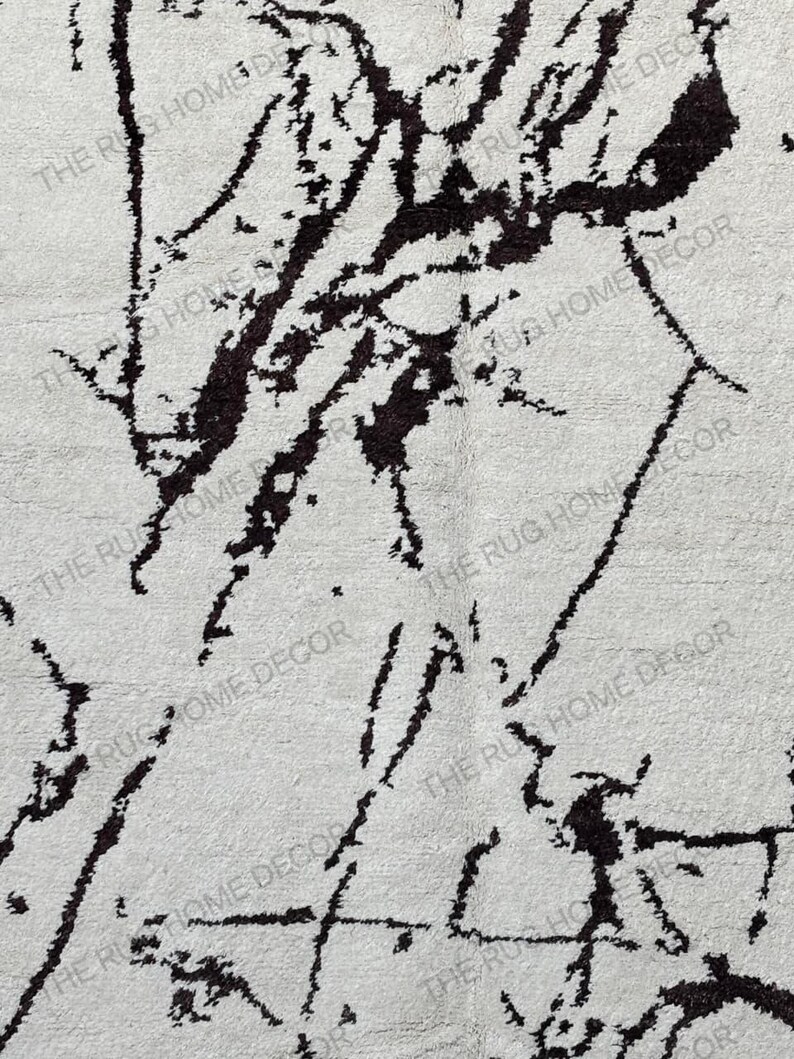 May include: A close-up of a white rug with a black abstract design. The rug has a textured surface and the words "THE RUG HOME DECOR" are printed on the rug.