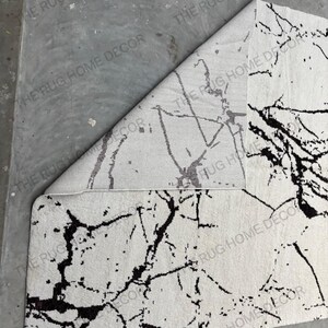May include: A white rug with a black marble-like pattern. The rug is partially rolled up, revealing the underside, which has a similar but less defined pattern. The rug's design is reminiscent of natural stone.
