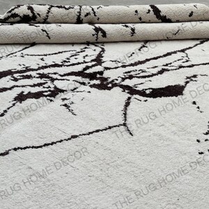 May include: A white rug with a dark brown, marble-like pattern. The rug is partially rolled up, revealing its textured surface. The words "THE RUG HOME DECOR" are printed on the rug.