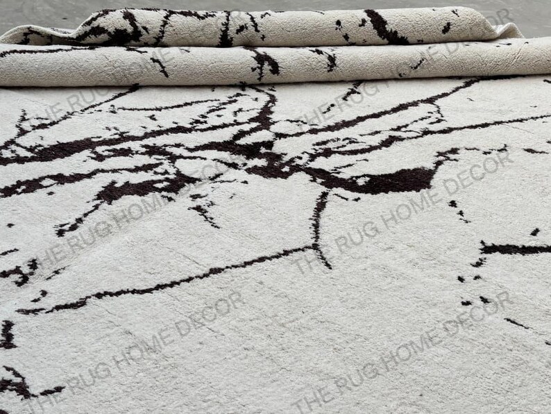 May include: A white rug with a dark brown, marble-like pattern. The rug is partially rolled up, revealing its textured surface. The words "THE RUG HOME DECOR" are printed on the rug.
