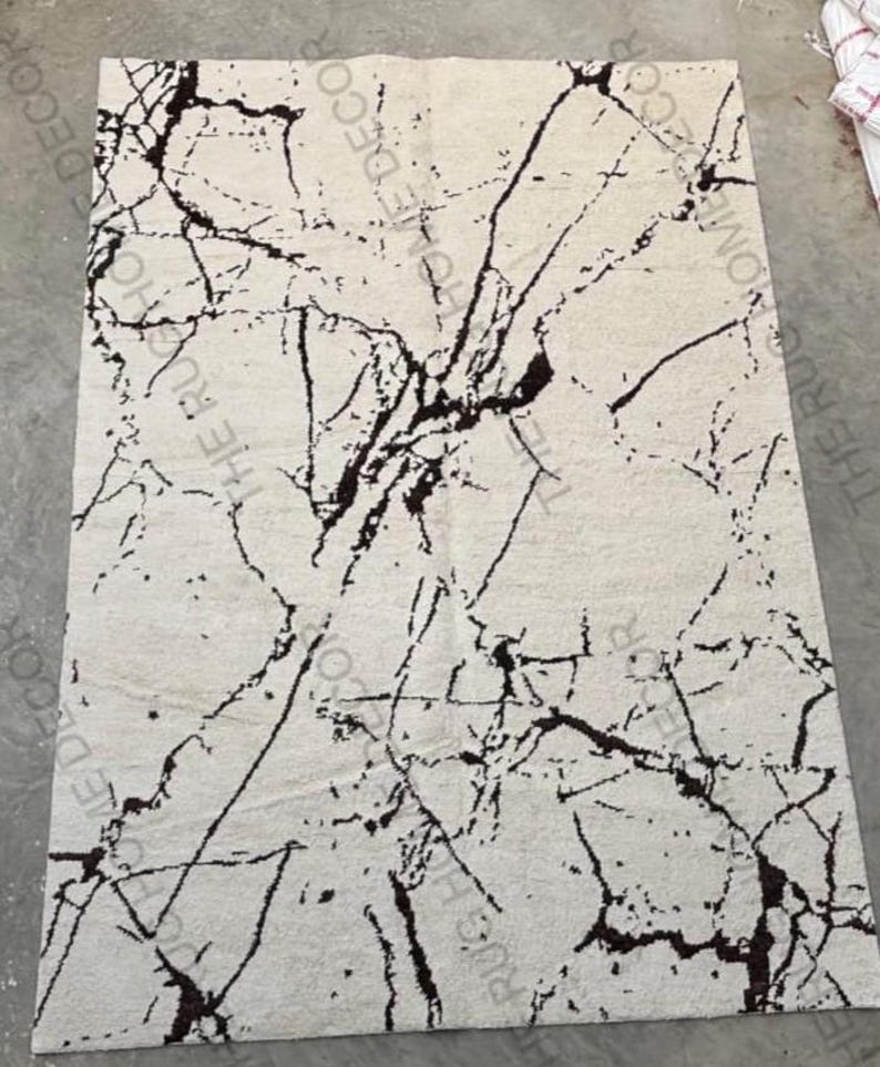 May include: A rectangular rug with a cream-colored background and a striking black marble-like pattern. The design features irregular, vein-like lines that create a dynamic visual texture. The rug is likely made of a soft material.