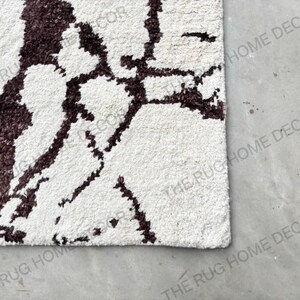 May include: Close-up of a white rug with a dark brown, abstract pattern. The rug has a textured surface and a soft appearance. The design is reminiscent of a natural, organic motif. The rug is a home decor item.