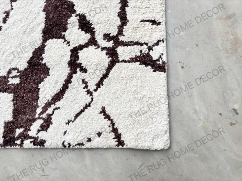May include: Close-up of a white rug with a dark brown, abstract pattern. The rug has a textured surface and a soft appearance. The design is reminiscent of a natural, organic motif. The rug is a home decor item.