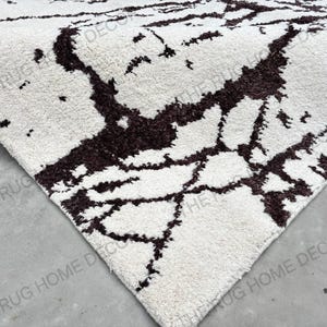 May include: Close-up of a white rug with a dark brown, abstract pattern. The rug's texture is visible, and the design resembles a network of lines. The corner of the rug is visible, showing its thickness. The words "THE RUG HOME DECOR" are printed on the rug.