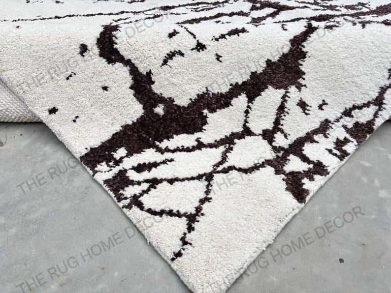 May include: Close-up of a white rug with a dark brown, abstract pattern. The rug's texture is visible, and the design resembles a network of lines. The corner of the rug is visible, showing its thickness. The words "THE RUG HOME DECOR" are printed on the rug.