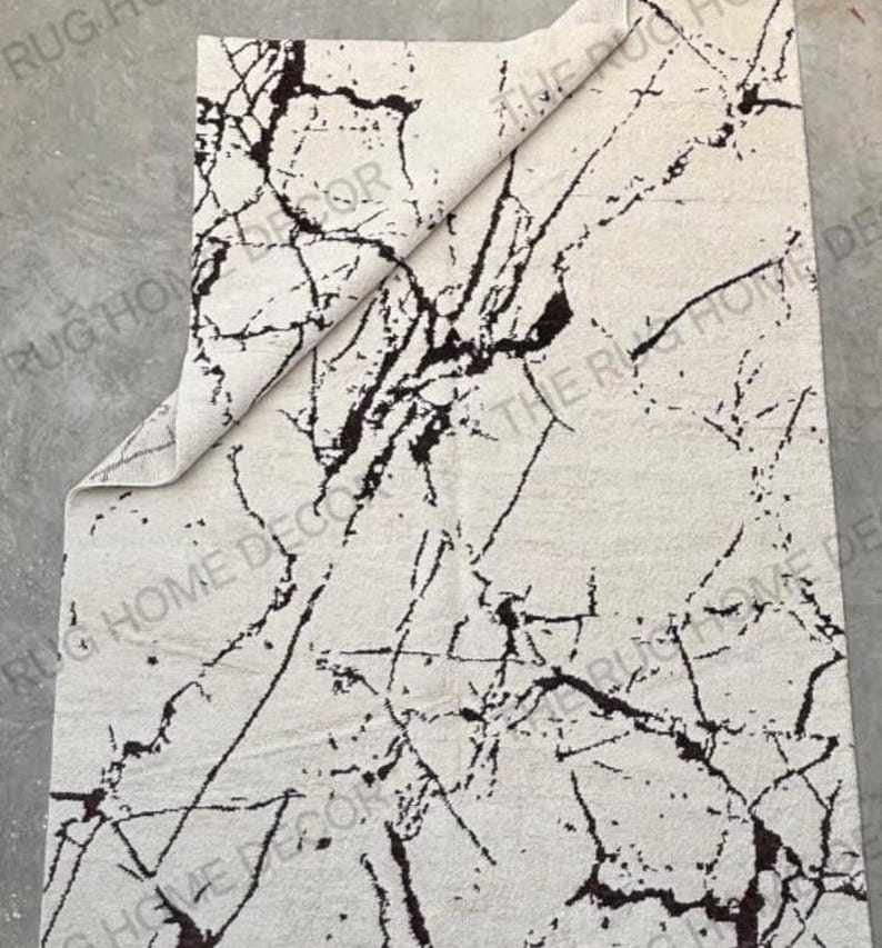 May include: A rectangular rug with a cream-colored background and a dark brown, marble-like pattern. The rug's corner is folded over, revealing the underside. The rug is designed for home decor.