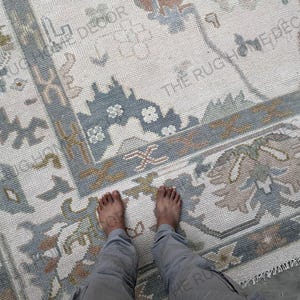 Op de afbeelding: Een rechthoekig vloerkleed met een crèmekleurige achtergrond en een gedetailleerde rand in tinten blauw, bruin en groen. Het vloerkleed heeft bloemen- en geometrische patronen. De afbeelding toont de voeten en benen van een persoon.