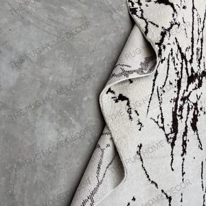 May include: A white rug with black abstract patterns, partially rolled up to reveal the underside. The rug has a textured surface and the text "THE RUG HOME DECOR" is visible.