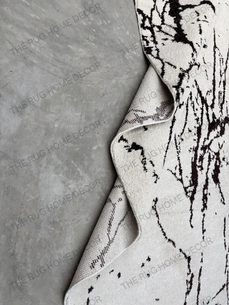May include: A white rug with black abstract patterns, partially rolled up to reveal the underside. The rug has a textured surface and the text "THE RUG HOME DECOR" is visible.