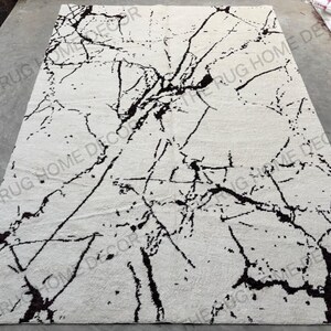 May include: A rectangular, off-white rug with a striking black marble-like pattern. The design features bold, irregular black lines and veins across the surface, creating a modern, artistic look. The rug is likely made of a soft, textured material.