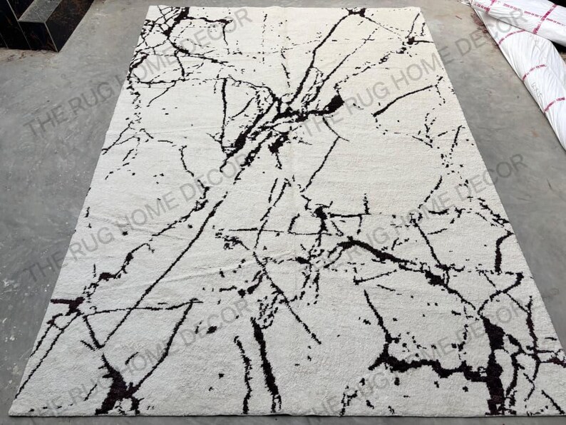 May include: A rectangular, off-white rug with a striking black marble-like pattern. The design features bold, irregular black lines and veins across the surface, creating a modern, artistic look. The rug is likely made of a soft, textured material.