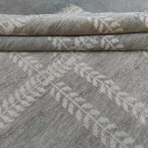 May include: A folded grey rug featuring a repeating white leaf design. The rug has a textured surface and a neutral colour scheme, suitable for various interior styles. The edges of the rug are visible, showing the woven construction.