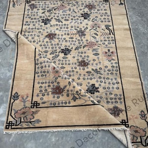 May include: Beige and brown area rug with a floral pattern and a black border. The rug features a central design of flowers and leaves in shades of brown, black, and pink. The rug is rectangular and has a folded corner.