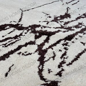 May include: Close-up of a cream-colored rug with a dark brown, abstract pattern. The rug has a soft, textured appearance, with the dark brown design resembling organic shapes. The words "THE RUG HOME DECOR" are printed on the rug.
