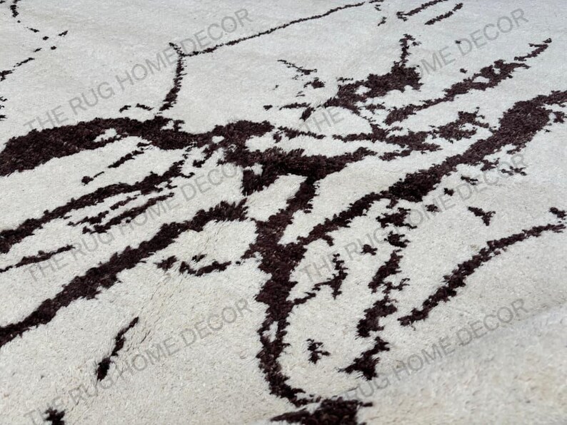 May include: Close-up of a cream-colored rug with a dark brown, abstract pattern. The rug has a soft, textured appearance, with the dark brown design resembling organic shapes. The words "THE RUG HOME DECOR" are printed on the rug.