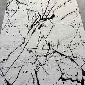 May include: A rectangular area rug with a white background and a black marble-like pattern. The rug features a network of dark, irregular lines resembling veins across its surface. The words "THE RUG HOME DECOR" are printed on the rug.