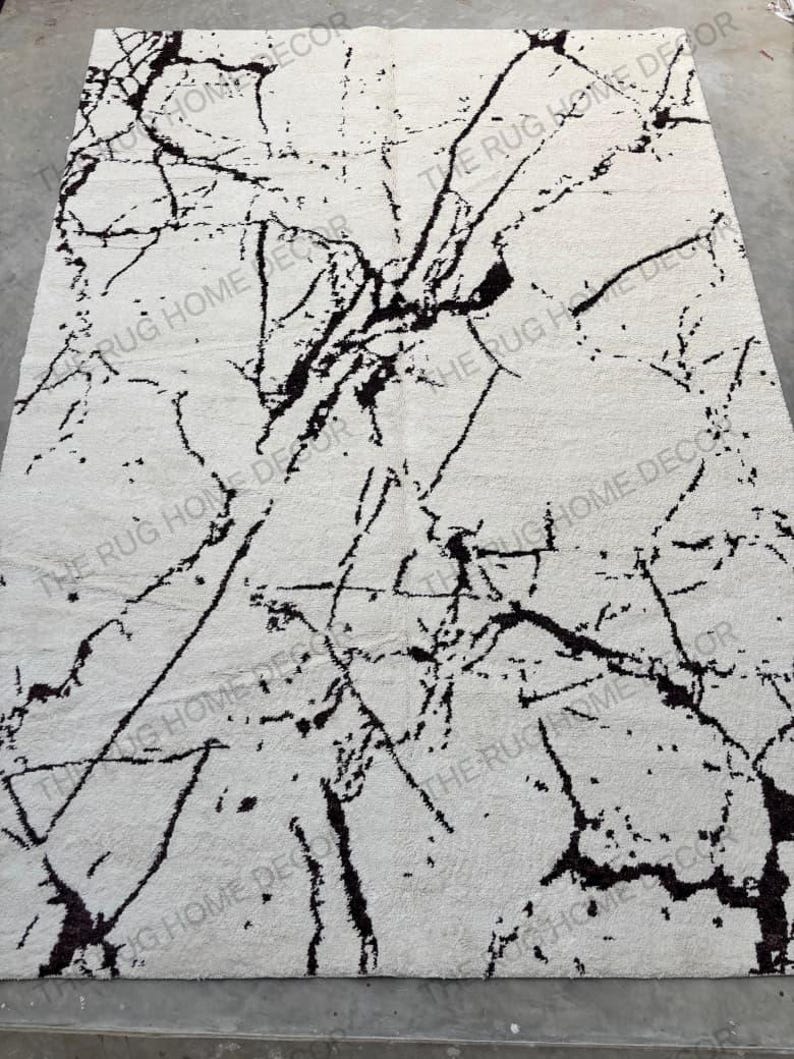 May include: A rectangular area rug with a white background and a black marble-like pattern. The rug features a network of dark, irregular lines resembling veins across its surface. The words "THE RUG HOME DECOR" are printed on the rug.
