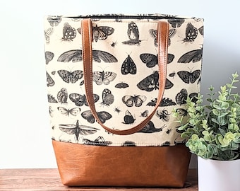 May include: A beige canvas tote bag with brown leather handles and a brown leather bottom. The bag is printed with a black and white pattern of butterflies.