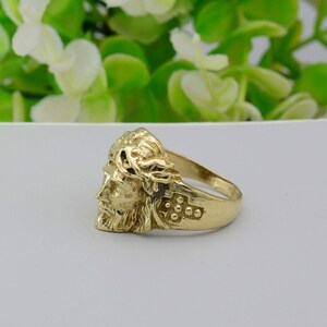 May include: A gold ring with a detailed depiction of Jesus's face wearing a crown of thorns. The ring also features a cross with small dots on the side.