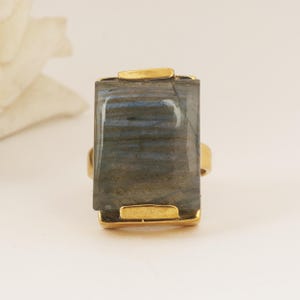 Puede incluir: Un anillo dorado con una piedra labradorita rectangular. La piedra presenta tonos grises, azules y verdes. La banda y el engaste del anillo son dorados, creando un contraste con los tonos naturales de la piedra.