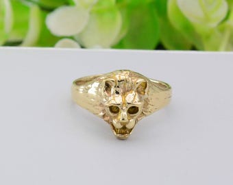 May include: A gold ring with a lion's head design. The ring has a textured surface and the lion's face is sculpted with visible eyes, nose, and mouth. The ring is set against a white background.