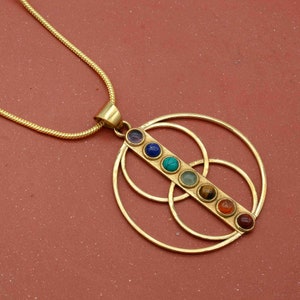 Könnte beinhalten: Eine goldene Halskette mit einem Anhänger, der sieben farbige Edelsteine ​​in einer vertikalen Linie innerhalb eines kreisförmigen Designs zeigt. Der Anhänger ist aus Messing und hat ein geometrisches Muster.