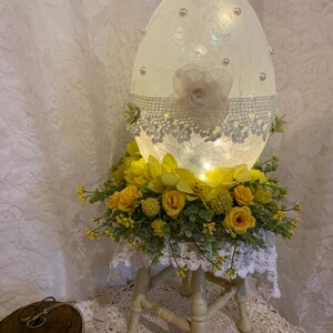 Puede incluir: Un gran objeto decorativo en forma de huevo iluminado. El huevo blanco presenta detalles de perlas, encaje y flores. Se apoya sobre un pequeño soporte de madera blanco, adornado con flores amarillas y vegetación.