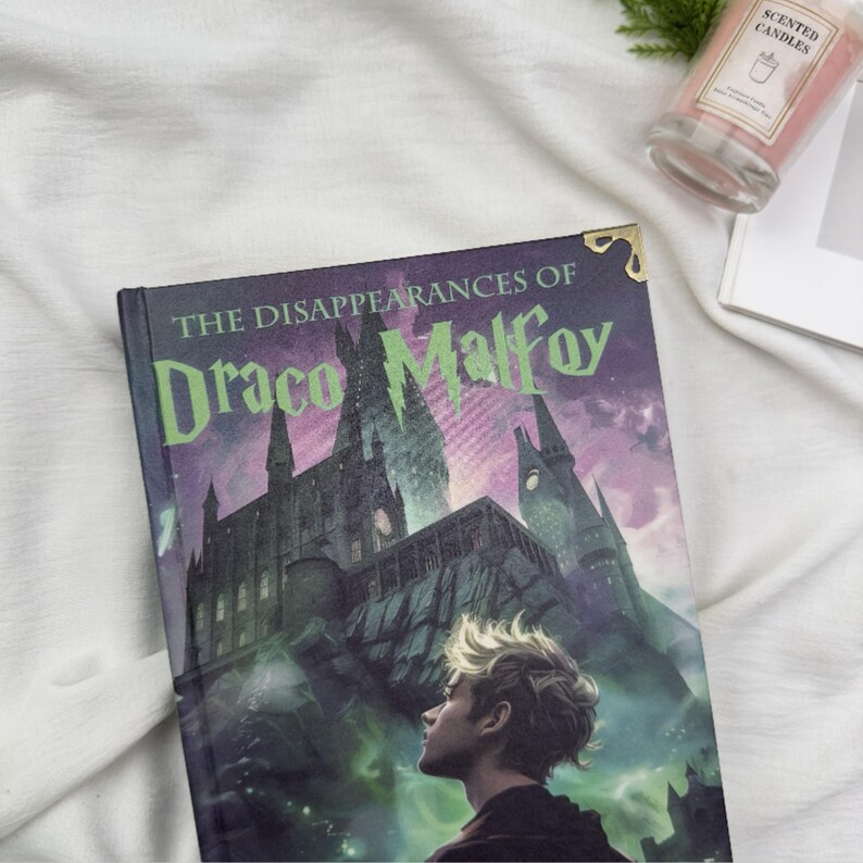 Puede incluir: Un libro de tapa dura titulado "The Disappearances of Draco Malfoy" con una ilustraci&oacute;n de un castillo y un retrato de un joven. El libro tiene una combinaci&oacute;n de colores p&uacute;rpura y verde. Una vela perfumada y un libro est&aacute;n en el fondo.