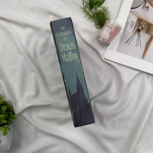 Puede incluir: Un libro titulado "The Disappearances of Draco Malfoy" con una cubierta azul oscuro y verde. La cubierta presenta una silueta de un castillo. El libro est&aacute; sobre un fondo blanco.