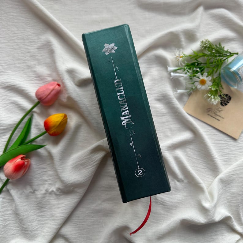 May include: A dark teal book with the title "MANACLED" in silver lettering. A white flower and stem design is above the title. A red ribbon bookmark extends from the bottom. The number "2" is at the bottom. The book is on a white cloth.