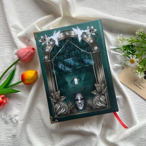 May include: A hardback book with a teal cover featuring an illustration of a building and the word "SAVOY". The cover also includes a silver mask, floral accents, and a red ribbon bookmark. Two white bird cutouts are at the top.