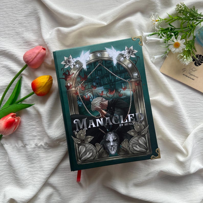 May include: Hardcover book titled "Manacled" by Senlinyu. The book has a dark teal cover with gold accents and a detailed illustration of a person and a mask. The cover art includes white angel figures and red origami cranes.