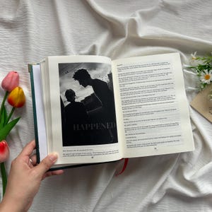 May include: An open book with a green cover and a black and white image that says "HAPPENED". The image shows two silhouetted figures. The book is held open by a hand, with text on the right page. Red ribbon bookmark.
