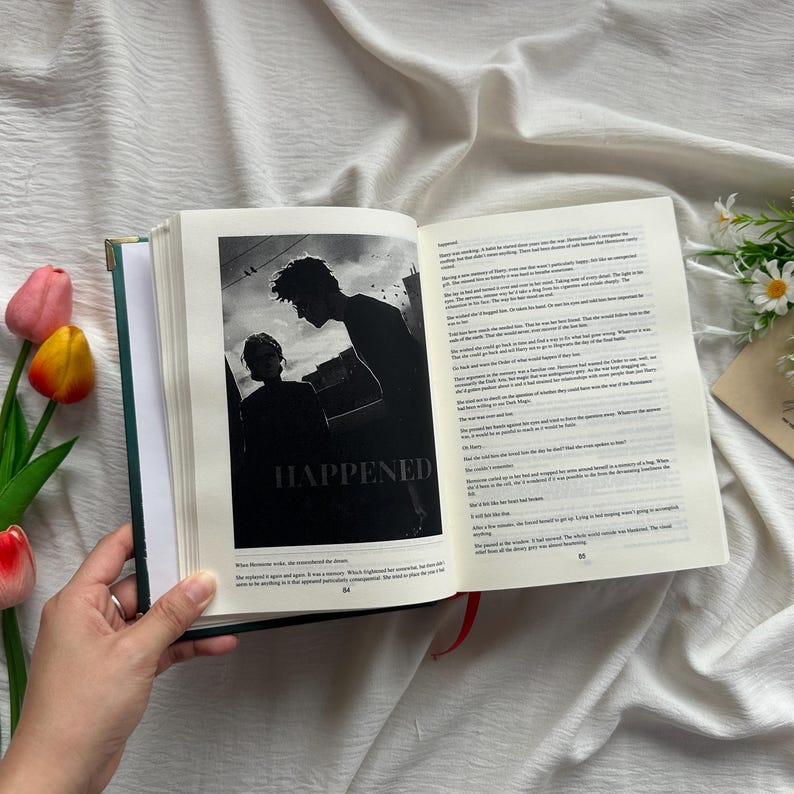 May include: An open book with a green cover and a black and white image that says "HAPPENED". The image shows two silhouetted figures. The book is held open by a hand, with text on the right page. Red ribbon bookmark.