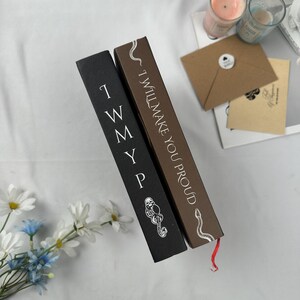 May include: Two hardcover books with text. One is black with white text reading "I W M Y P" and a skull and snake graphic. The other is brown with the text "I WILL MAKE YOU PROUD" in white. A red ribbon bookmark is visible.