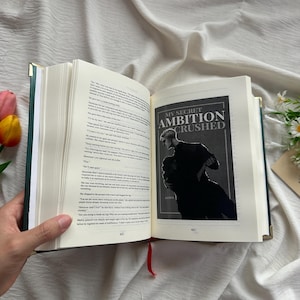 May include: An open book with a green cover and gold-colored edges, revealing printed pages and a book cover illustration. The cover illustration features a black and white image with the text "MY SECRET AMBITION CRUSHED". A red ribbon bookmark is visible.
