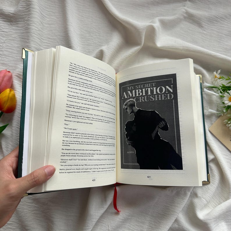 May include: An open book with a green cover and gold-colored edges, revealing printed pages and a book cover illustration. The cover illustration features a black and white image with the text "MY SECRET AMBITION CRUSHED". A red ribbon bookmark is visible.