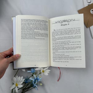 May include: An open book with text on the pages, featuring a gold-edged cover and a red ribbon bookmark. The text includes the chapter title "Chapter 6". Artificial blue and white flowers are in the foreground.