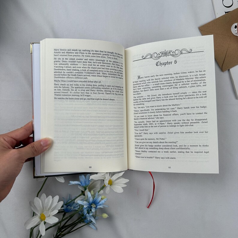 May include: An open book with text on the pages, featuring a gold-edged cover and a red ribbon bookmark. The text includes the chapter title "Chapter 6". Artificial blue and white flowers are in the foreground.