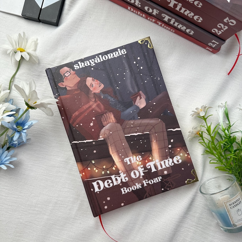 May include: A hardcover book titled "The Debt of Time, Book Four" with an illustration of a couple. The cover features the author's name, "shay&aacute;lonnie," and a red bookmark. Other books are visible in the background.