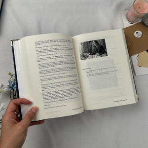 May include: An open book with text and a black and white image. The book has a navy blue cover with gold edges. A hand is holding the book open to a page with text and an image of a person.